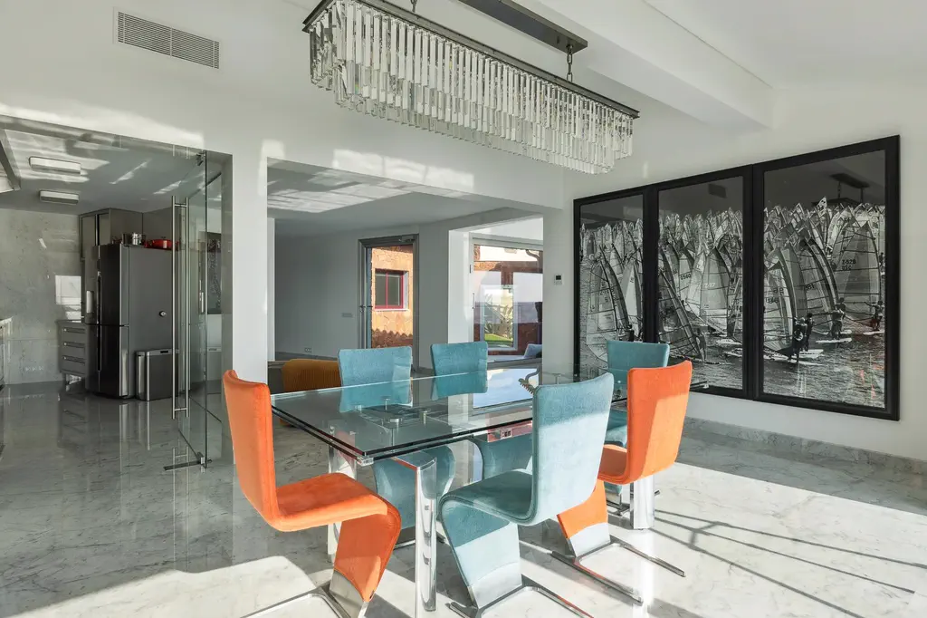 La salle à manger, chaises orange et table de verre, la cuisine derrière la paroi vitrée